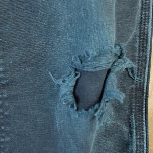 May include: Close-up of distressed blue denim jeans. The jeans have a large, frayed hole, revealing the fabric underneath. The stitching and texture of the denim are visible, with a light-colored wooden surface in the background.