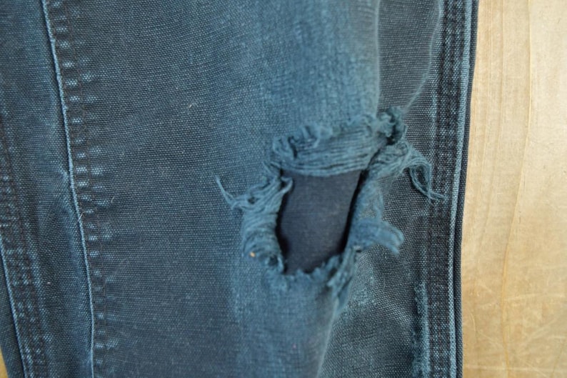 May include: Close-up of distressed blue denim jeans. The jeans have a large, frayed hole, revealing the fabric underneath. The stitching and texture of the denim are visible, with a light-colored wooden surface in the background.