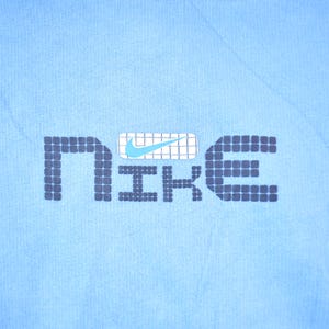 May include: A light blue t-shirt with a black and white pixelated "NIKE" logo. The logo has a blue swoosh inside a white box.