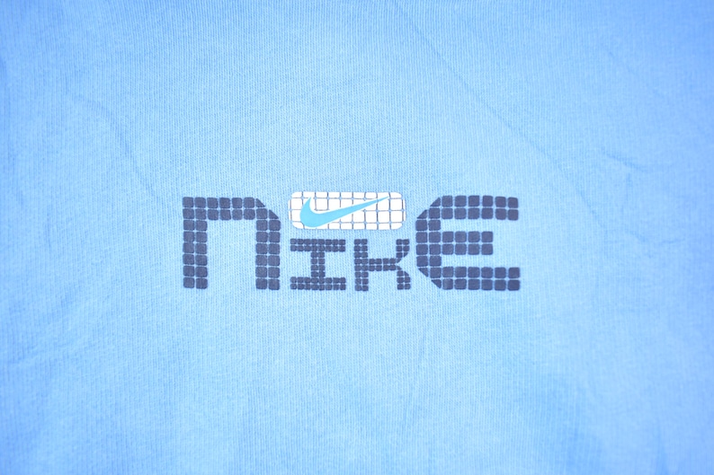 May include: A light blue t-shirt with a black and white pixelated "NIKE" logo. The logo has a blue swoosh inside a white box.