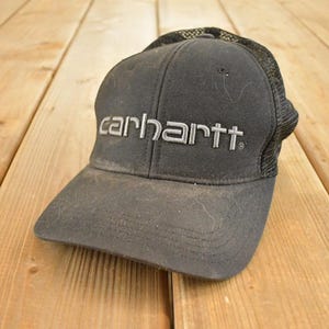 Puede incluir: Una gorra de béisbol Carhartt negra con la parte trasera de malla. El logotipo de Carhartt está bordado en gris en la parte delantera de la gorra. La gorra está sobre una superficie de madera.