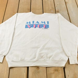Könnte beinhalten: Weißes Sweatshirt mit Rundhalsausschnitt und dem Text "MIAMI VICE" in blauen und rosa Buchstaben. Das Sweatshirt hat lange Ärmel und gerippte Bündchen und Saum. Das Kleidungsstück ist aus einem weichen, bequemen Material.