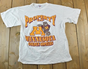 May include: White t-shirt with a graphic featuring the text "PROPERTY OF MINNESOTA GOLDEN GOPHERS" in orange and purple. A cartoon gopher wearing a maroon and gold jersey is in the center. The shirt is laid flat on a wooden surface.
