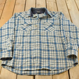 May include: A long-sleeved, button-down shirt with a blue and white plaid pattern. The shirt has two chest pockets and a dark blue collar. The cuffs are buttoned. The shirt is lying on a wooden surface.