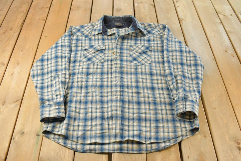 May include: A long-sleeved, button-down shirt with a blue and white plaid pattern. The shirt has two chest pockets and a dark blue collar. The cuffs are buttoned. The shirt is lying on a wooden surface.