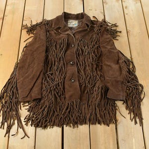 May include: Brown suede jacket with fringe detailing. The jacket has a collared neckline, button-up front, and long fringe along the sleeves, chest, and hem. A vintage-style piece.
