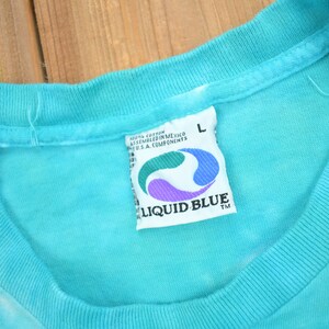 Puede incluir: Una camiseta azul turquesa con una etiqueta de Liquid Blue. La etiqueta es blanca con un dise&ntilde;o de remolino azul y morado. La etiqueta dice "Liquid Blue" y "100% algod&oacute;n ensamblado en M&eacute;xico Componentes de EE. UU.". La camiseta es talla L.