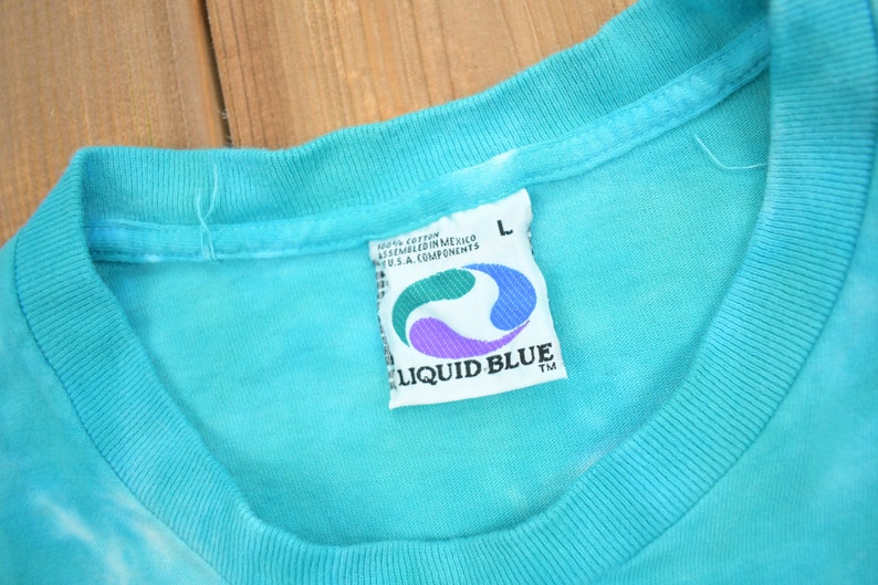 Puede incluir: Una camiseta azul turquesa con una etiqueta de Liquid Blue. La etiqueta es blanca con un dise&ntilde;o de remolino azul y morado. La etiqueta dice "Liquid Blue" y "100% algod&oacute;n ensamblado en M&eacute;xico Componentes de EE. UU.". La camiseta es talla L.
