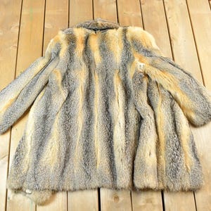 May include: A vintage fur coat with a striped pattern in shades of gray and tan. The coat has long sleeves and a collar. The fur appears soft and plush, with a natural texture. The coat is laid out on a wooden surface.