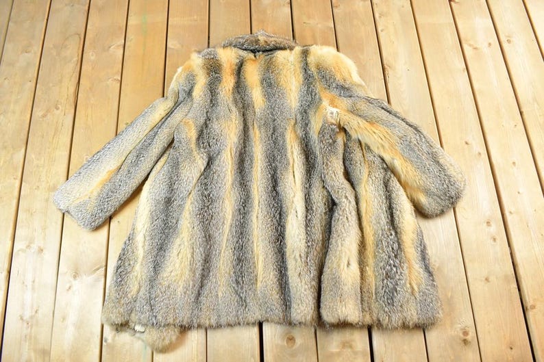 May include: A vintage fur coat with a striped pattern in shades of gray and tan. The coat has long sleeves and a collar. The fur appears soft and plush, with a natural texture. The coat is laid out on a wooden surface.