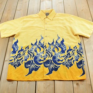 May include: A short-sleeved button-down shirt in a bright yellow color. The lower portion of the shirt features a bold, repeating graphic pattern of stylized blue flames and floral shapes that extend upward from the hem toward the chest area.