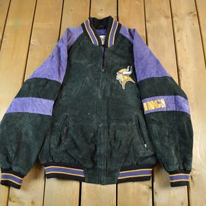 Op de afbeelding: Een vintage zwarte en paarse suède jas met ritssluiting. De jas heeft paarse mouwen met zwarte strepen en het woord "VINGS" op de mouw. Een geel en paars logo staat op de borst.