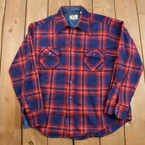 Puede incluir: Una camisa de franela abotonada de manga larga con un clásico estampado a cuadros rojo y azul marino. La camisa tiene dos bolsillos en el pecho y un cuello. Los puños y el cuello son de un azul más oscuro. La camisa está sobre una superficie de madera.