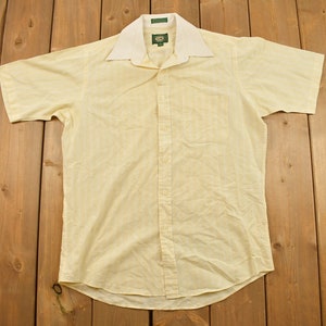 Puede incluir: Una camisa amarilla de manga corta con botones y cuello blanco. La camisa tiene un patrón sutil y un solo bolsillo en el pecho izquierdo.