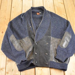 May include: A navy blue knit cardigan with leather accents. The sweater features a shawl collar, leather patches on the elbows, and a double-breasted front with silver buttons. The leather is a dark gray color.