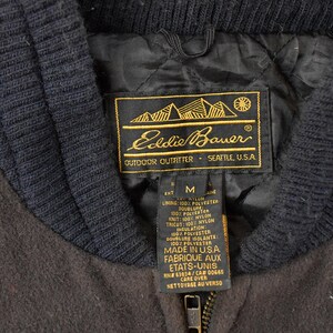 May include: A dark brown jacket with a black ribbed collar and a black quilted interior lining. The Eddie Bauer label is stitched inside, featuring gold text and a mountain graphic, along with a size M tag and care instructions in English and French.