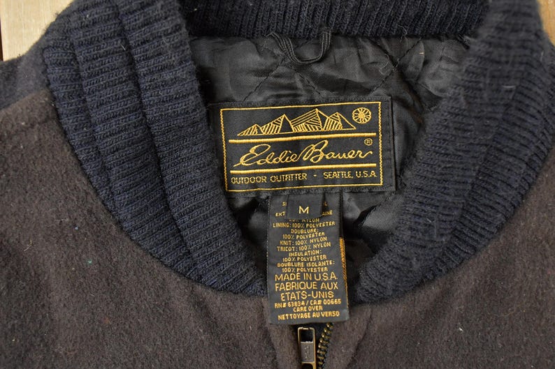 May include: A dark brown jacket with a black ribbed collar and a black quilted interior lining. The Eddie Bauer label is stitched inside, featuring gold text and a mountain graphic, along with a size M tag and care instructions in English and French.