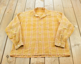 May include: A long-sleeve button-down shirt in a mustard yellow hue featuring a plaid pattern with thin white and red vertical and horizontal lines. The shirt includes a pointed collar, a single chest pocket, and white buttons on the front placket.