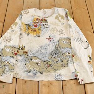 May include: Cream-colored long-sleeve shirt featuring a world map design with illustrations of buildings, snowflakes, and Santa Claus. The map includes compass roses and nautical charts. The shirt is laid flat on a wooden surface.