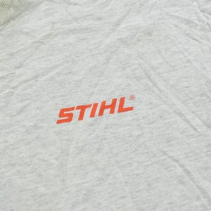 Op de afbeelding: Een lichtgrijs gem&ecirc;leerd T-shirt met het oranje STIHL-logo op de borst. De stof heeft een textuur met een gespikkeld uiterlijk en een geribbelde ronde hals die zichtbaar is in de linkerbovenhoek van het kledingstuk.