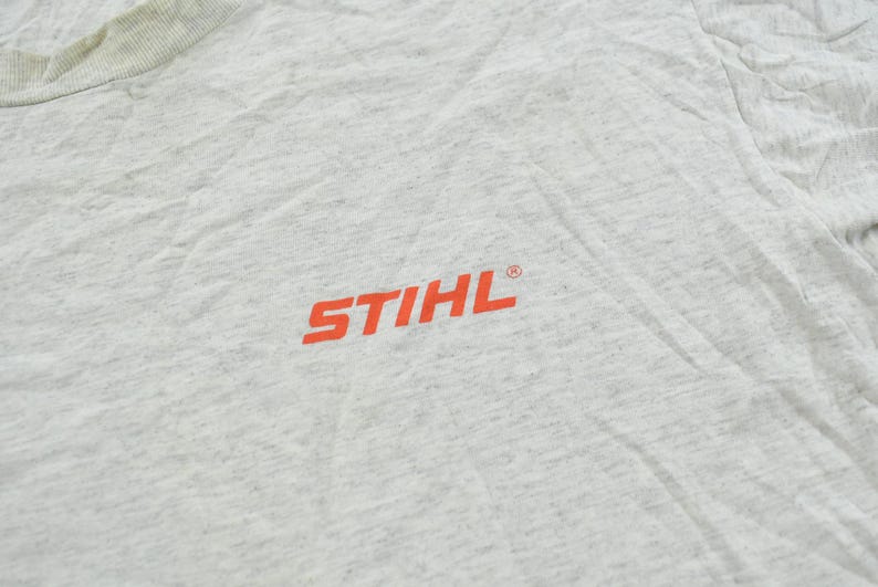 Op de afbeelding: Een lichtgrijs gem&ecirc;leerd T-shirt met het oranje STIHL-logo op de borst. De stof heeft een textuur met een gespikkeld uiterlijk en een geribbelde ronde hals die zichtbaar is in de linkerbovenhoek van het kledingstuk.