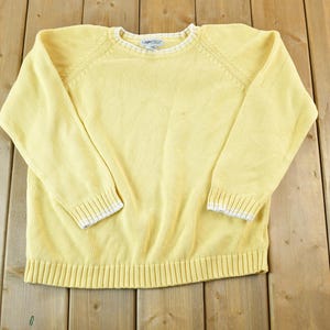 Puede incluir: Un suéter amarillo claro con cuello redondo y mangas largas. Los puños y el cuello tienen un ribete blanco. El suéter está hecho de tejido de punto y tiene un dobladillo acanalado. El suéter está sobre una superficie de madera.