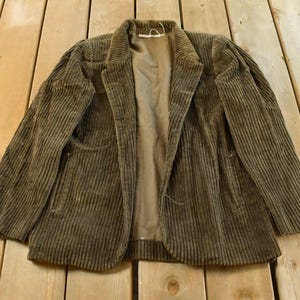 Puede incluir: Blazer vintage de pana verde oliva con solapas de muesca y dos bolsillos delanteros. La chaqueta está abierta, revelando un forro beige. La tela de pana tiene una textura acanalada vertical. El blazer está sobre una superficie de madera.