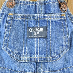 May include: Close-up of blue denim overalls with the "OshKosh B'gosh" logo on the pocket. The overalls have copper-colored buttons and white stitching. The text "THE GENUINE ARTICLE" is below the logo.