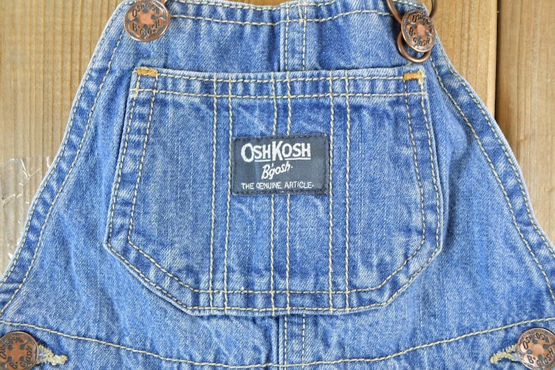 May include: Close-up of blue denim overalls with the "OshKosh B'gosh" logo on the pocket. The overalls have copper-colored buttons and white stitching. The text "THE GENUINE ARTICLE" is below the logo.