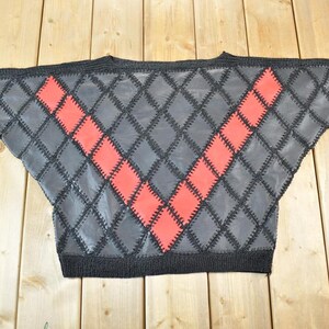 Könnte beinhalten: Ein grauer Pullover mit Rautenmuster und einem roten Zickzack-Design. Der Pullover hat einen schwarzen Strickkragen, Manschetten und Saum. Das Rautenmuster ist mit schwarzem Faden genäht.