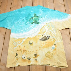 Puede incluir: Una camiseta tie-dye con una escena de playa que presenta una tortuga marina, un p&aacute;jaro y conchas marinas sobre un fondo amarillo.