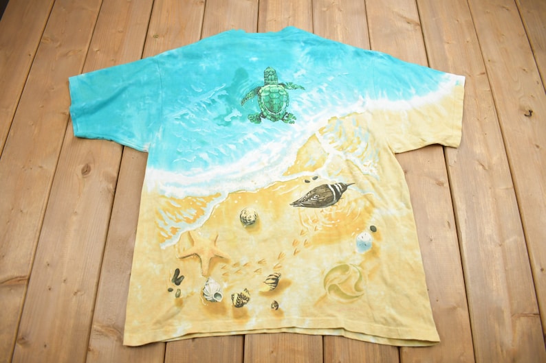 Puede incluir: Una camiseta tie-dye con una escena de playa que presenta una tortuga marina, un p&aacute;jaro y conchas marinas sobre un fondo amarillo.