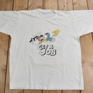 May include: White t-shirt featuring a cartoon illustration of a character falling and the text "GET A JOB".