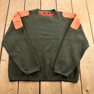 May include: A dark green sweater with orange elbow patches. The sweater has a crew neck and long sleeves.