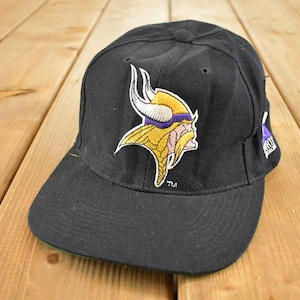 Può includere: Cappellino da baseball nero con il logo dei Minnesota Vikings. Il logo è una testa di vichingo in giallo, viola e bianco. Il cappellino ha una visiera curva e un lato inferiore verde.