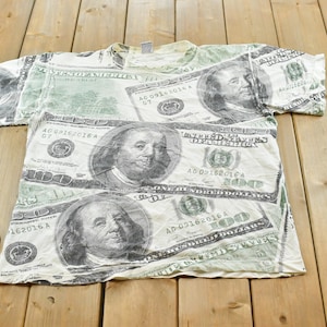 May include: A short-sleeved t-shirt featuring an all-over print of United States one hundred dollar bills. The design includes the portrait of Benjamin Franklin, the text One Hundred Dollars, and the United States of America printed in green and white.