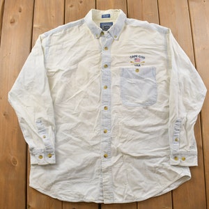 Op de afbeelding: Een wit overhemd met lange mouwen en knopen met een blauwe denim zak. Het shirt heeft een geborduurd logo met de tekst "Cape Cod USA" met een kleine Amerikaanse vlag.