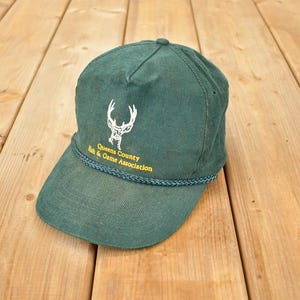 Può includere: Cappellino da baseball in velluto a coste verde con visiera curva. Il cappellino presenta un emblema di cervo bianco e la scritta "Queens County Fish & Game Association" in giallo. Un dettaglio di corda intrecciata adorna la visiera.