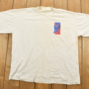 Peut inclure: T-shirt blanc avec un motif graphique bleu et rouge sur le devant. Le graphique représente une image stylisée d'une personne avec un microphone.