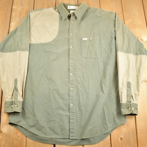 Puede incluir: Una camisa verde de manga larga con botones, un parche acolchado beige en el hombro y mangas beiges. La camisa tiene un solo bolsillo en el pecho.