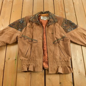 Könnte beinhalten: Eine braune Lederjacke mit Reißverschluss und Kordelzug in der Taille. Die Jacke hat einen gemusterten Stoffkragen und Schulterpartien. Das Etikett auf der Jacke lautet "Northgate Leatherwear".