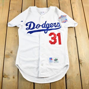 Puede incluir: Camiseta de béisbol blanca con la palabra "Dodgers" en escritura azul en el pecho y el número "31" en rojo. La camiseta tiene una parte delantera abotonada y un parche en la manga.