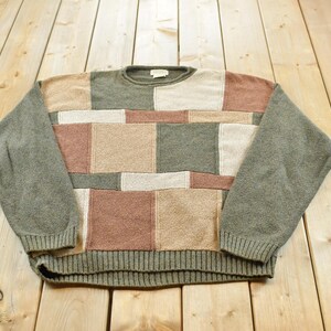 Puede incluir: Un suéter de manga larga con cuello redondo que presenta un diseño de parches con bloques rectangulares y cuadrados en tonos apagados de verde oliva, beige, crema y rosa viejo. La prenda tiene cuello, puños y dobladillo de punto acanalado.