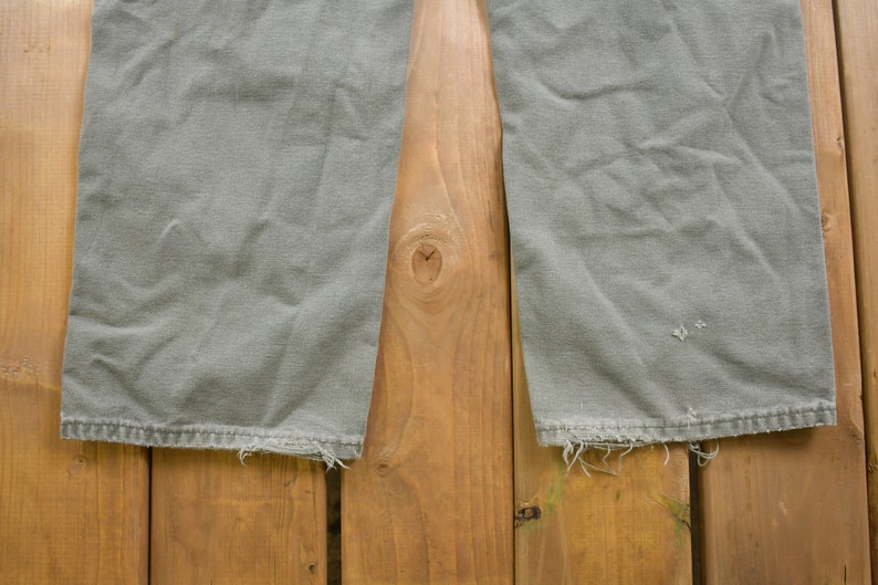 May include: A pair of grey cargo trousers with frayed hems. The trousers are laid flat on a wooden surface.