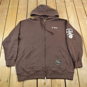 Op de afbeelding: Een bruine hoodie met rits en capuchon. De hoodie heeft een wit logo op de linkerborst met de tekst "G Unit" en een wit logo op de rechtermouw met de tekst "Property of G Unit".