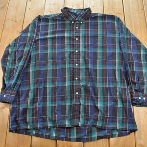 May include: A long-sleeve button-down shirt featuring a plaid pattern in shades of dark blue, forest green, and black with thin white and red vertical and horizontal lines. The shirt has a standard collar, a front button placket, and cuffs.