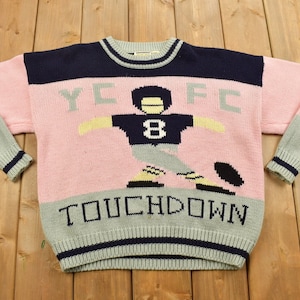 Op de afbeelding: Roze, grijze en donkerblauwe gebreide trui met een voetbalspeler afbeelding en de tekst "YCF" en "TOUCHDOWN".