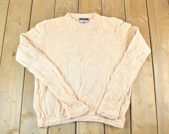 May include: A light peach-colored sweater with a textured pattern. The long-sleeved sweater has a crew neck and a ribbed hem and cuffs. The garment is laid flat on a wooden surface.