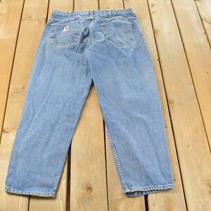 Puede incluir: Vaqueros de mezclilla azul claro con un dise&ntilde;o cl&aacute;sico de cinco bolsillos. Los vaqueros est&aacute;n colocados sobre una superficie de madera, mostrando los bolsillos traseros y las trabillas. Los vaqueros tienen un aspecto descolorido.