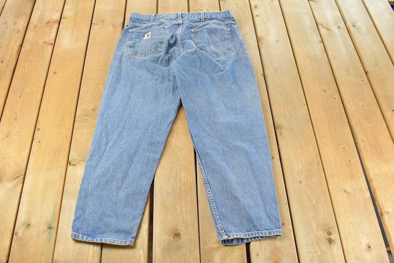 Puede incluir: Vaqueros de mezclilla azul claro con un dise&ntilde;o cl&aacute;sico de cinco bolsillos. Los vaqueros est&aacute;n colocados sobre una superficie de madera, mostrando los bolsillos traseros y las trabillas. Los vaqueros tienen un aspecto descolorido.